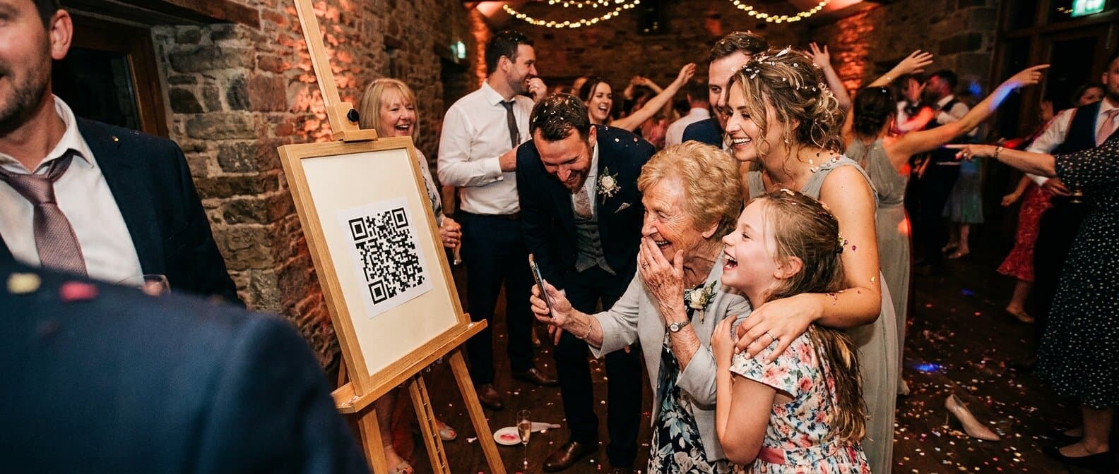 Lachende bruiloftsgasten van verschillende leeftijden scannen samen de QR-code op een bord naast de dansvloer